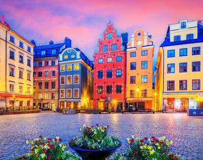Colorful historic buildings line a cobblestone square at sunset, with vibrant skies and blooming flowers in the foreground, creating a warm, inviting scene.