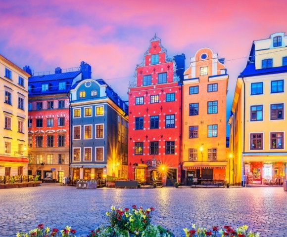 Colorful historic buildings line a cobblestone square at sunset, with vibrant skies and blooming flowers in the foreground, creating a warm, inviting scene.