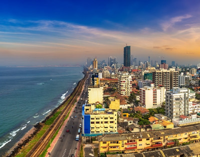 Aerial view of a vibrant coastal cityscape at sunset, featuring tall buildings lining the shoreline, a road along the coast, and the ocean under a colorful sky.