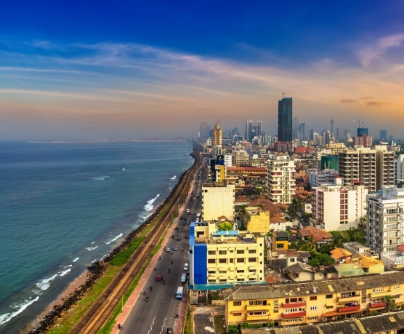 Aerial view of a vibrant coastal cityscape at sunset, featuring tall buildings lining the shoreline, a road along the coast, and the ocean under a colorful sky.