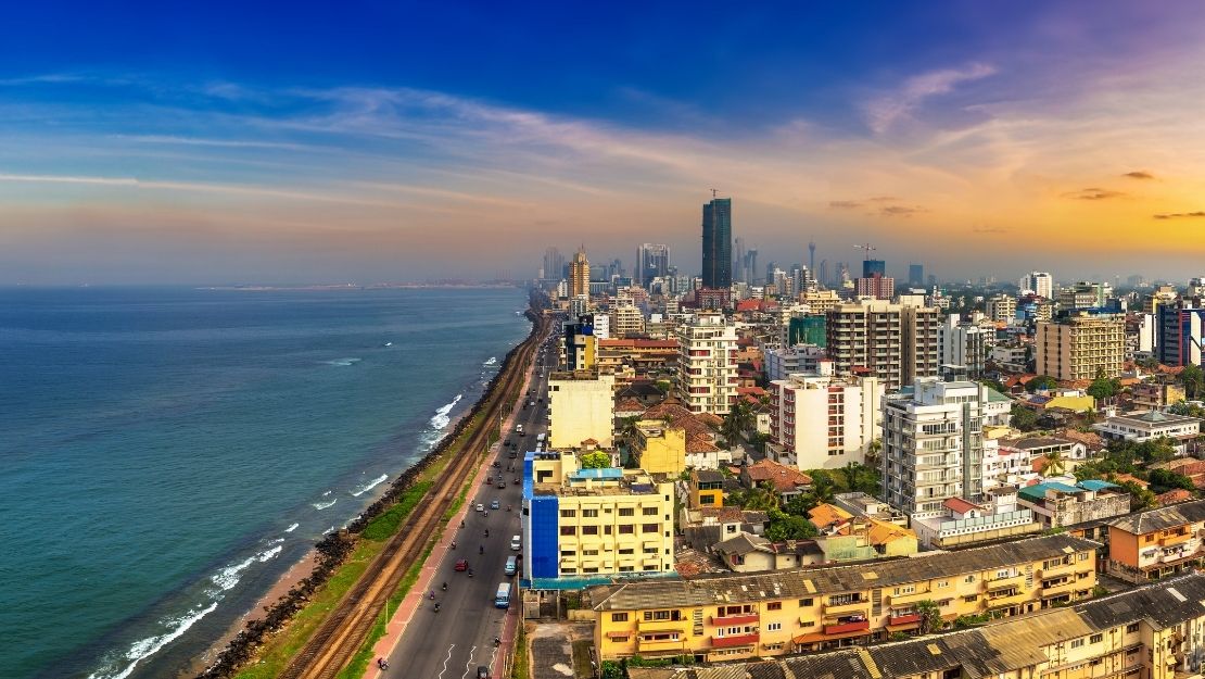 Aerial view of a vibrant coastal cityscape at sunset, featuring tall buildings lining the shoreline, a road along the coast, and the ocean under a colorful sky.