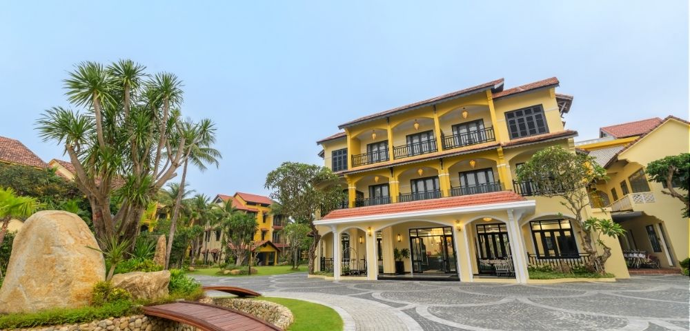 A serene view of a yellow colonial-style hotel with arched entrances, surrounded by palm trees and lush greenery under a clear blue sky.