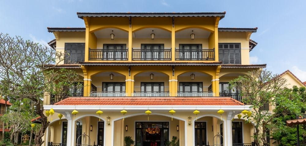 Three-story yellow building with arched balconies and black railings. Surrounded by lush greenery and blue sky, conveying a peaceful and inviting atmosphere.