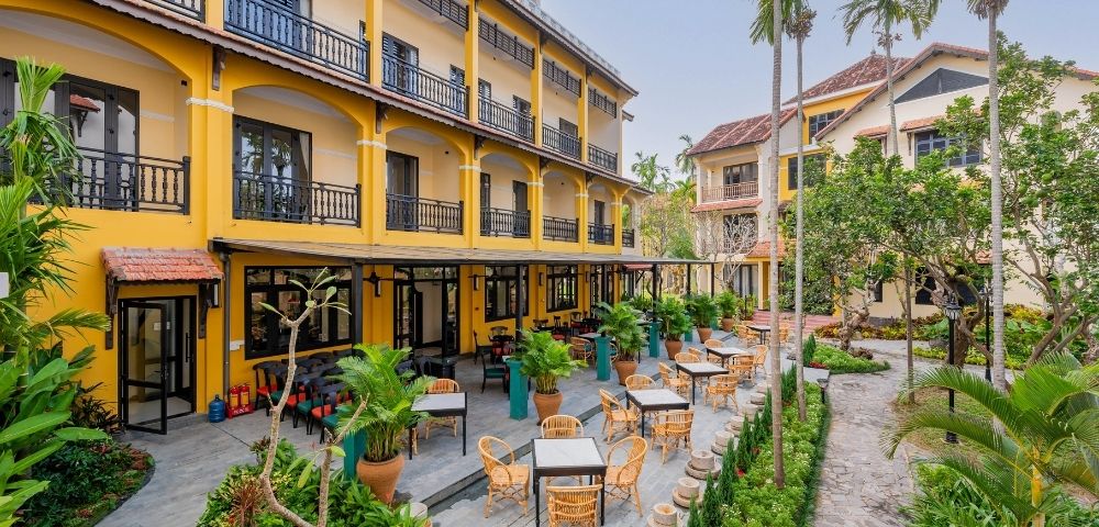 Courtyard seating area with outdoor tables and chairs in front of a yellow hotel building with balconies and lush greenery.