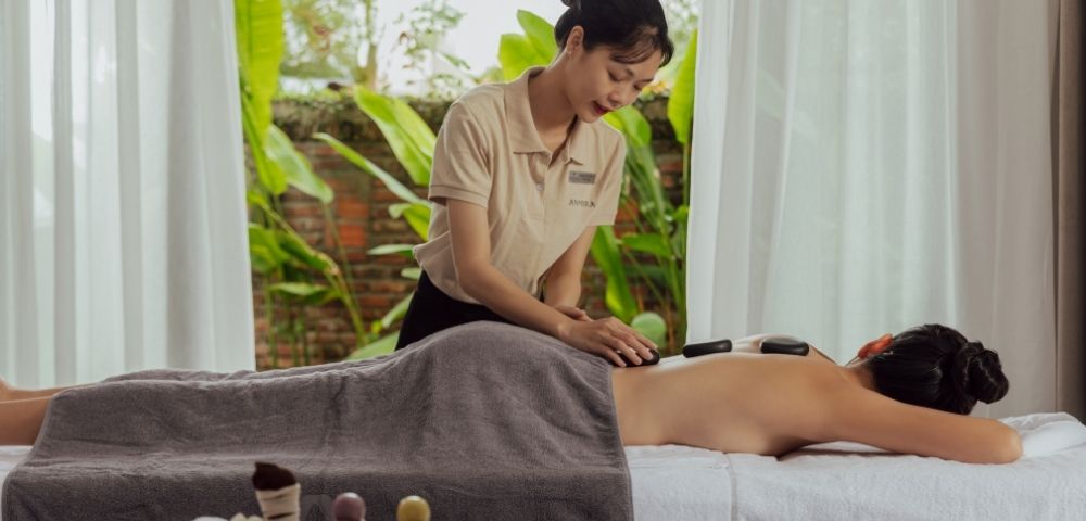 A spa therapist performs a hot stone massage on a client lying face down on a massage table.