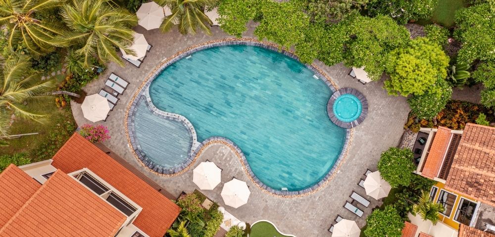 Aerial view of a resort with a large, irregularly shaped swimming pool and a separate circular jacuzzi, surrounded by lush green trees, palm trees, lounge chairs, and buildings with orange tiled roofs.