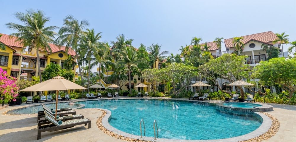 A resort swimming pool with turquoise water, surrounded by lounge chairs, umbrellas, tropical palm trees, and buildings with terracotta roofs under a clear blue sky.