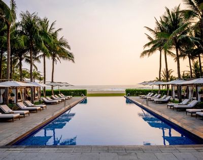 A serene infinity pool faces a tranquil ocean at sunset, flanked by rows of white lounge chairs, cabanas, and tall palm trees against a warm, gradient sky.
