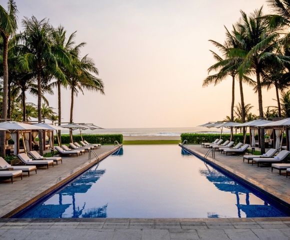 A serene infinity pool faces a tranquil ocean at sunset, flanked by rows of white lounge chairs, cabanas, and tall palm trees against a warm, gradient sky.