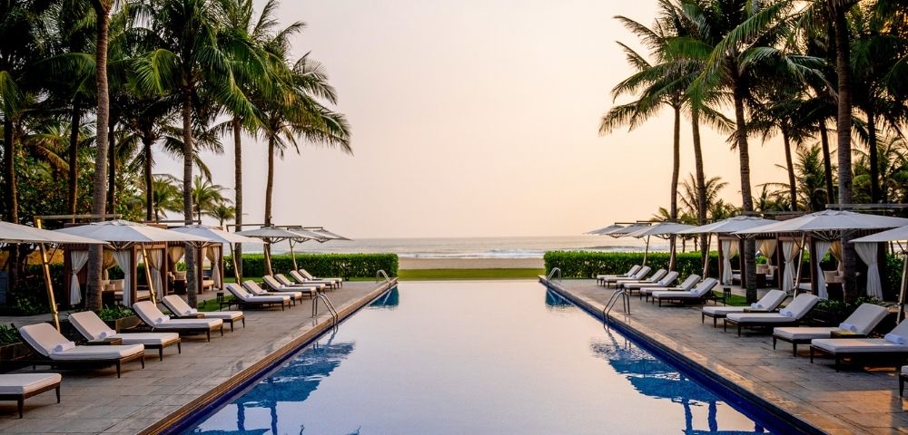 A serene infinity pool faces a tranquil ocean at sunset, flanked by rows of white lounge chairs, cabanas, and tall palm trees against a warm, gradient sky.