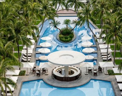 Aerial view of a resort swimming pool area with palm trees, lounge chairs, umbrellas, and a circular bar.