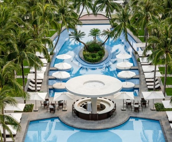 Aerial view of a resort swimming pool area with palm trees, lounge chairs, umbrellas, and a circular bar.