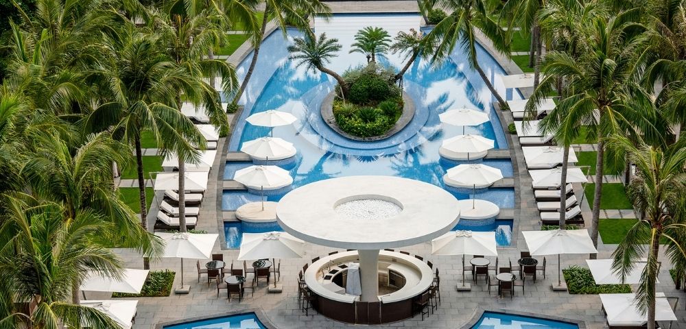 Aerial view of a resort swimming pool area with palm trees, lounge chairs, umbrellas, and a circular bar.