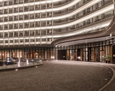 Modern hotel entrance at night with illuminated curved facade, outdoor fountain, and a paved driveway.