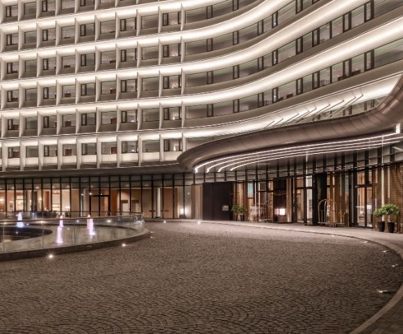 Modern hotel entrance at night with illuminated curved facade, outdoor fountain, and a paved driveway.