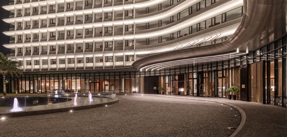 Modern hotel entrance at night with illuminated curved facade, outdoor fountain, and a paved driveway.