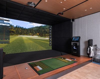 A high-tech indoor golf simulator setup with a large screen displaying a virtual golf course and performance data, a hitting mat with a golf ball, a golf bag, and a collection of golf clubs.