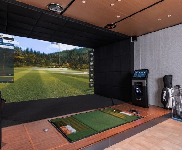 A high-tech indoor golf simulator setup with a large screen displaying a virtual golf course and performance data, a hitting mat with a golf ball, a golf bag, and a collection of golf clubs.