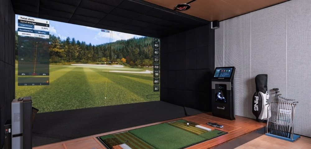 A high-tech indoor golf simulator setup with a large screen displaying a virtual golf course and performance data, a hitting mat with a golf ball, a golf bag, and a collection of golf clubs.
