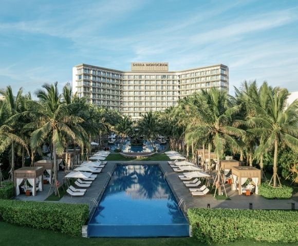 A large resort complex with a curved, modern hotel building, palm trees, lush greenery, and multiple swimming pools and cabanas under a bright blue sky.