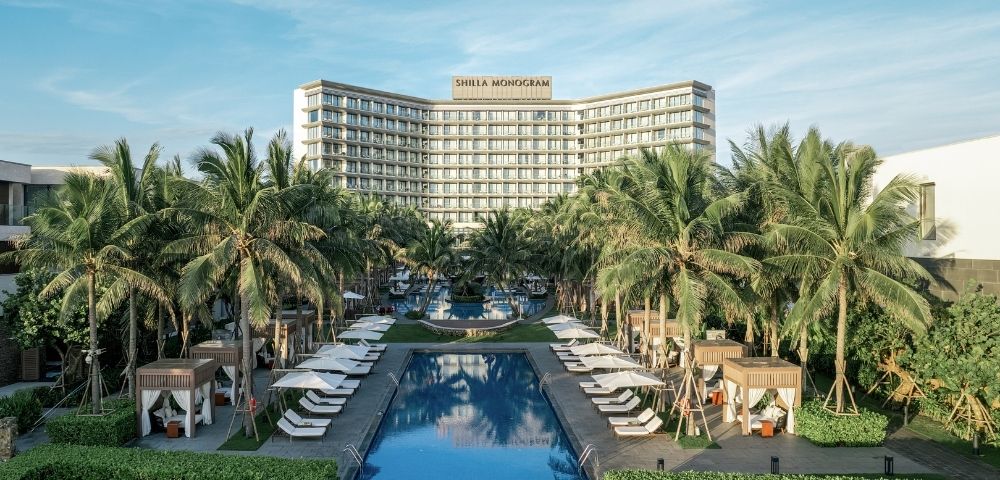 A large resort complex with a curved, modern hotel building, palm trees, lush greenery, and multiple swimming pools and cabanas under a bright blue sky.