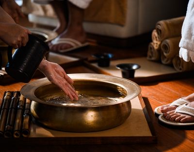 Hands pour water from a dark pitcher into a brass basin filled with clear liquid, with bamboo sticks arranged to the left and feet in flip-flops to the right.