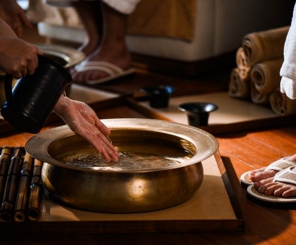 Hands pour water from a dark pitcher into a brass basin filled with clear liquid, with bamboo sticks arranged to the left and feet in flip-flops to the right.