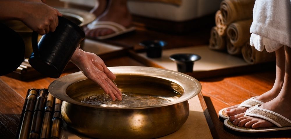 Hands pour water from a dark pitcher into a brass basin filled with clear liquid, with bamboo sticks arranged to the left and feet in flip-flops to the right.