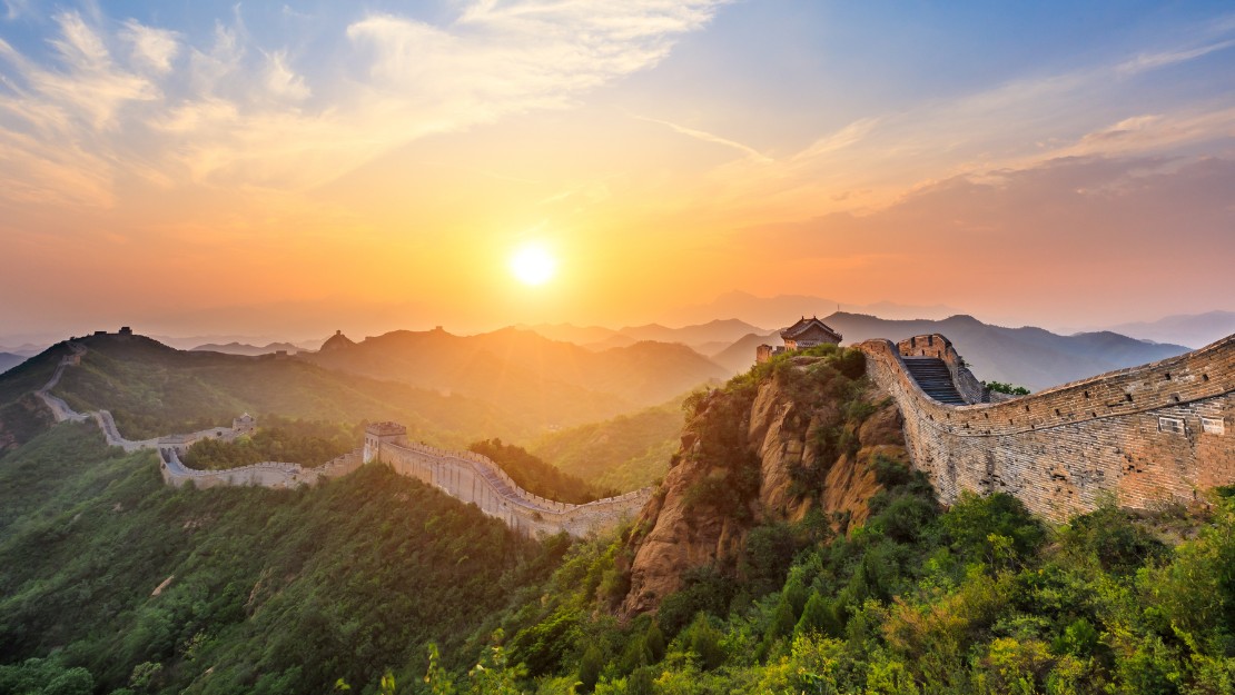 Sunset over the Great Wall of China, with lush green hills and a vibrant sky. The scene evokes a sense of majesty and tranquility.