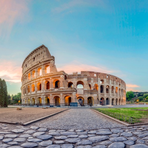 Ancient Roman Colosseum at sunset, with its iconic arches illuminated. A stone pathway leads to the structure, and the sky is a vibrant blue and pink.