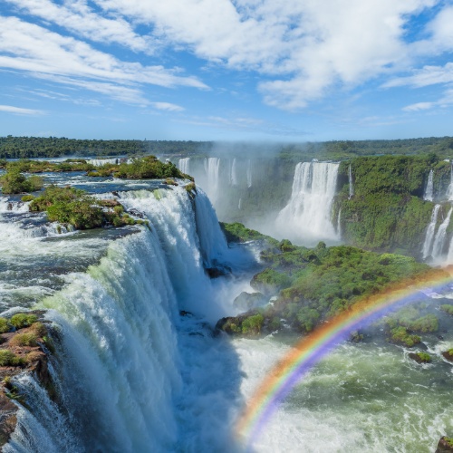 A majestic waterfall cascades over lush green cliffs under a partly cloudy sky, with a vibrant rainbow arching across the scene, creating a serene atmosphere.