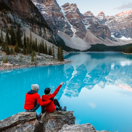 Two people in red jackets sit on rocks by a serene blue lake, surrounded by pine trees and snowy mountains, with one person pointing skyward.