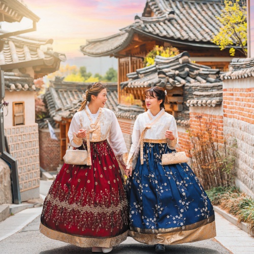 Two women in traditional Korean hanboks, one red and one blue, walk joyfully through a historical village with tiled roofs at sunset.