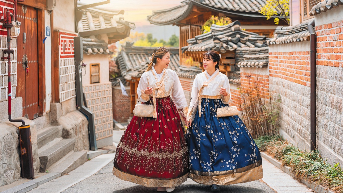Two women in traditional Korean hanboks, one red and one blue, walk joyfully through a historical village with tiled roofs at sunset.