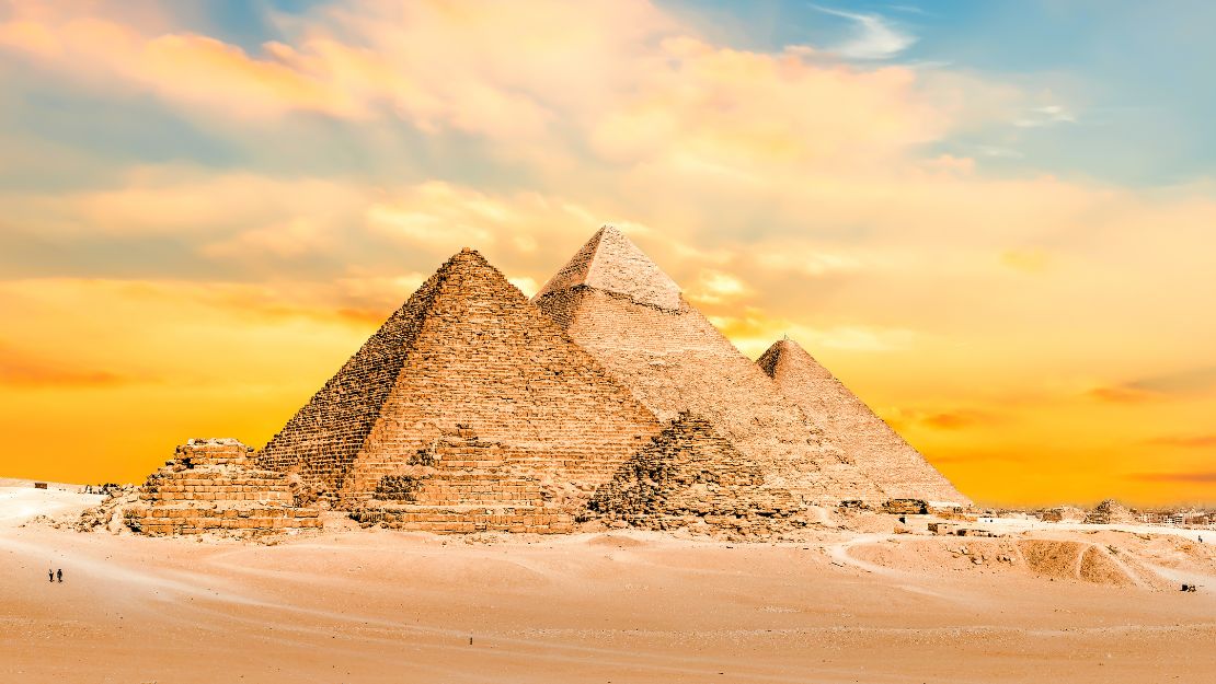The image shows the iconic pyramids of Giza under a vibrant, golden sunset sky. The desert foreground enhances their ancient, majestic presence.