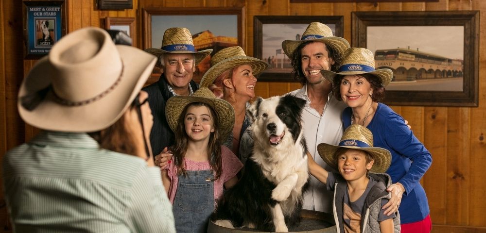 A diverse group of people and a border collie dog, all wearing straw hats, pose for a photo indoors against a wood-paneled wall with a vintage hotel photograph.