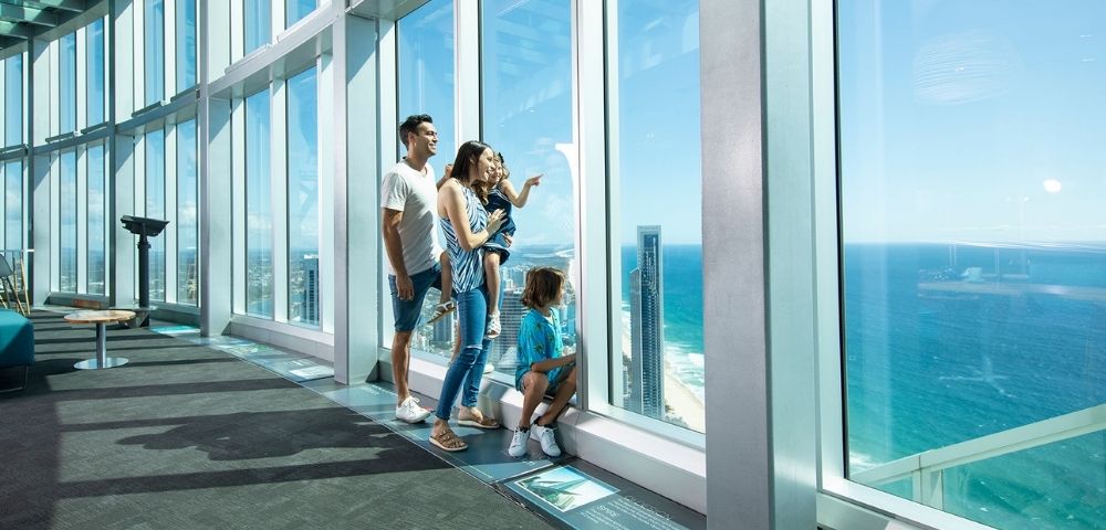 A family of four looks out of a large window from a high vantage point, with the ocean and a coastal city visible below.
