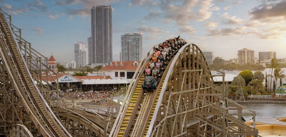 A wooden roller coaster with multiple cars full of passengers crests a hill, with a park and city skyline in the background.