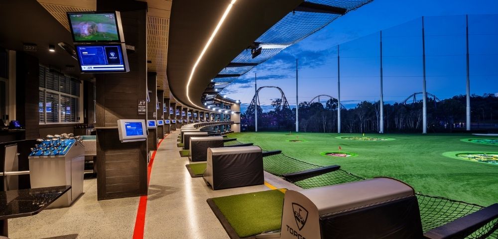 Panoramic view of a modern driving range at dusk, with rows of bays for hitting golf balls on the left and a large, illuminated green field with targets on the right, against a twilight sky with roller coaster structures in the background.