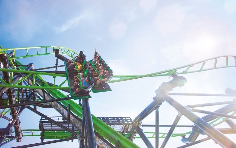 Two roller coaster trains with passengers ascend a steep green track against a bright blue sky with lens flare.