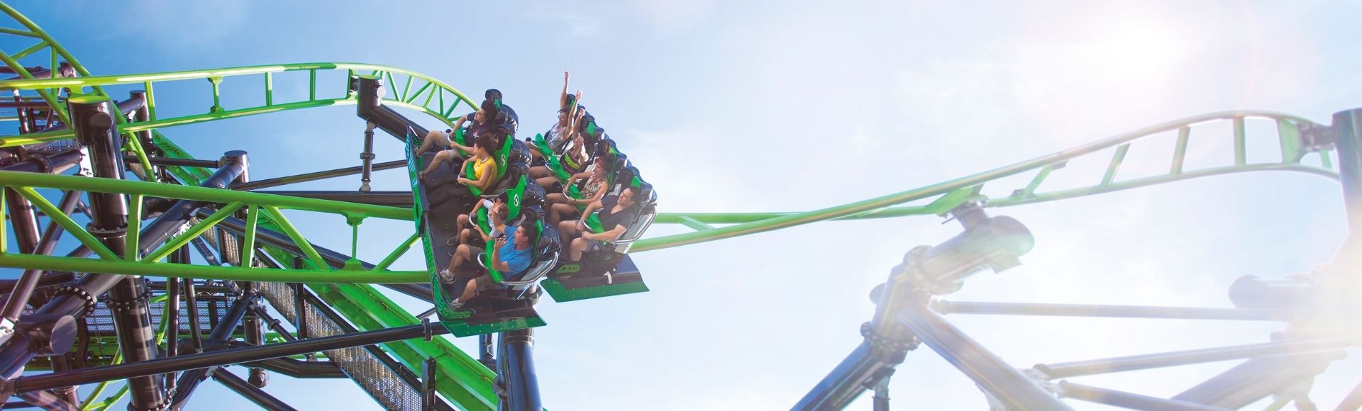 Two roller coaster trains with passengers ascend a steep green track against a bright blue sky with lens flare.