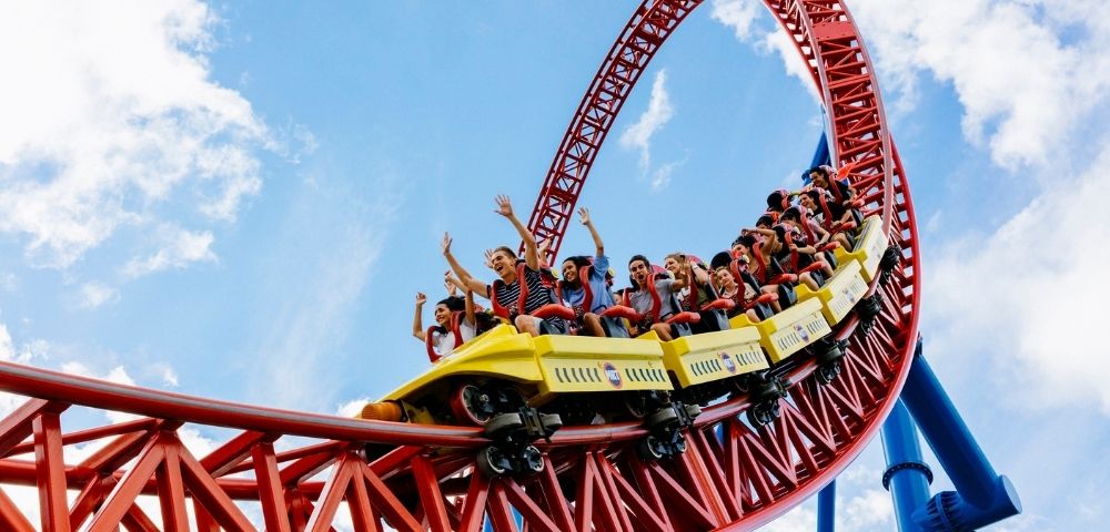 A bright yellow roller coaster car filled with excited passengers with hands raised in the air races along a steep red track against a vibrant blue sky with white clouds.