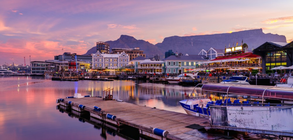 Scenic view of a harbor at sunset with vibrant sky, calm water, boats docked, and buildings lit up. Mountains in the background provide a serene backdrop.