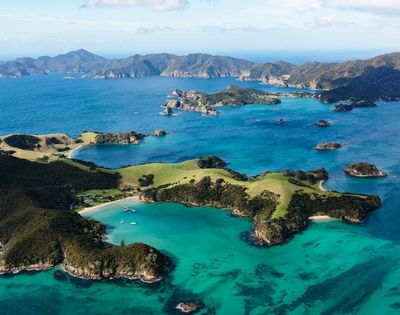Aerial view of a vibrant turquoise bay surrounded by lush green islands and rugged hills under a partly cloudy sky.