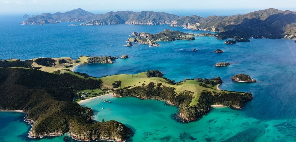 Aerial view of a vibrant turquoise bay surrounded by lush green islands and rugged hills under a partly cloudy sky.