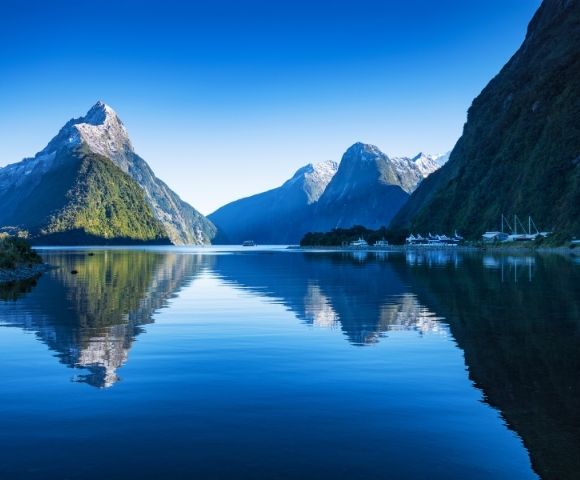 Calm blue lake reflecting towering green mountains under a clear sky, with boats docked near the shore.