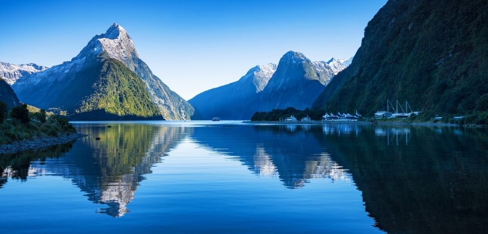 Calm blue lake reflecting towering green mountains under a clear sky, with boats docked near the shore.