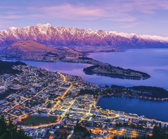 Aerial view of a lakeside town at dusk with glowing streetlights, a large island, and snow-capped mountains under a purple sky.