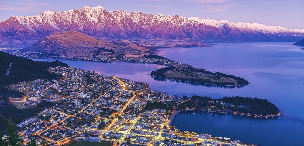 Aerial view of a lakeside town at dusk with glowing streetlights, a large island, and snow-capped mountains under a purple sky.