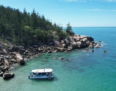 A white tour boat with blue accents floats in crystal clear turquoise water near a rocky, tree-covered island coastline under a bright blue sky.